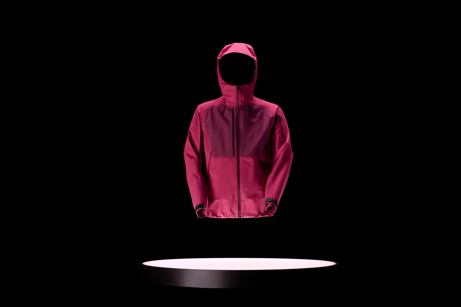 Rosa Sperro SV-jacka från Arc’teryx svävar över en rund belyst plattform mot svart bakgrund.
